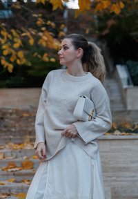 Bežani pleteni pulover s rebrastim manžetama, uparen s bijelom satenskom suknjom s čipkastim rubom. Drži malu bijelu torbicu s zlatnim akcentima.