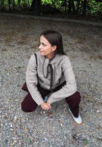 Kvinna med brunt hår som hukar på en grusväg, iförd en beige stickad tröja, mörka byxor, ljusa sneakers och med en väskrem över axeln.