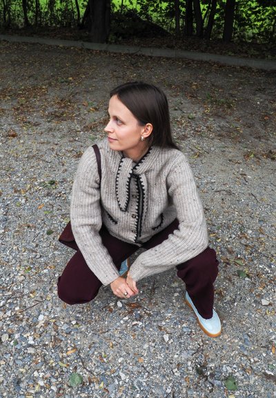 Mujer con cabello castaño agachada en un camino de grava, vistiendo un suéter de punto beige, pantalones oscuros, zapatillas claras, con una correa de bolsa sobre el hombro.
