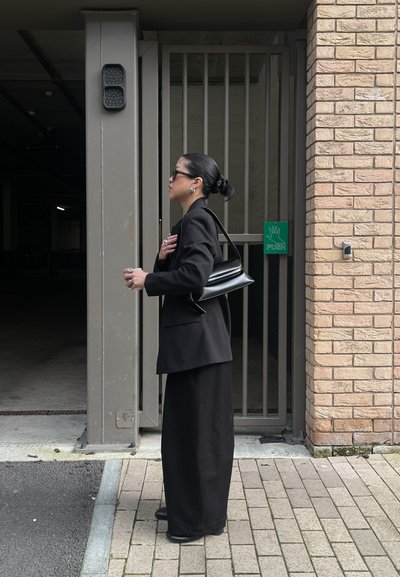 Mujer vestida con ropa negra y gafas de sol, lleva un bolso negro y está junto a una puerta metálica cerrada con un letrero verde que dice "EMPÚJALO".