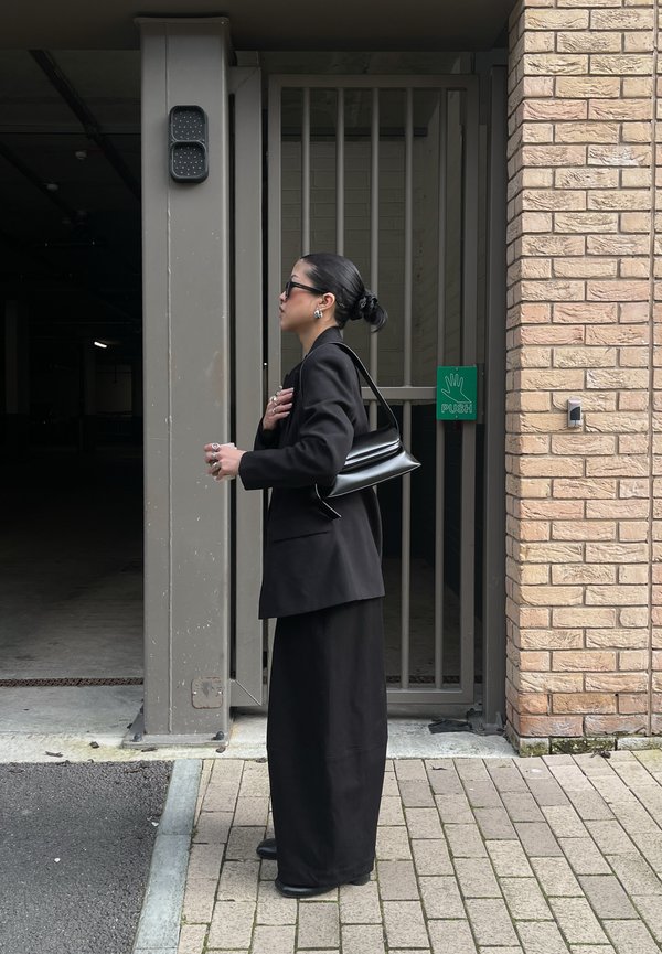 Žena v čiernom oblečení a slnečných okuliaroch, nesúca čiernu kabelku, stojí vedľa zatvorených kovových dverí so zeleným nápisom "TLAČ".