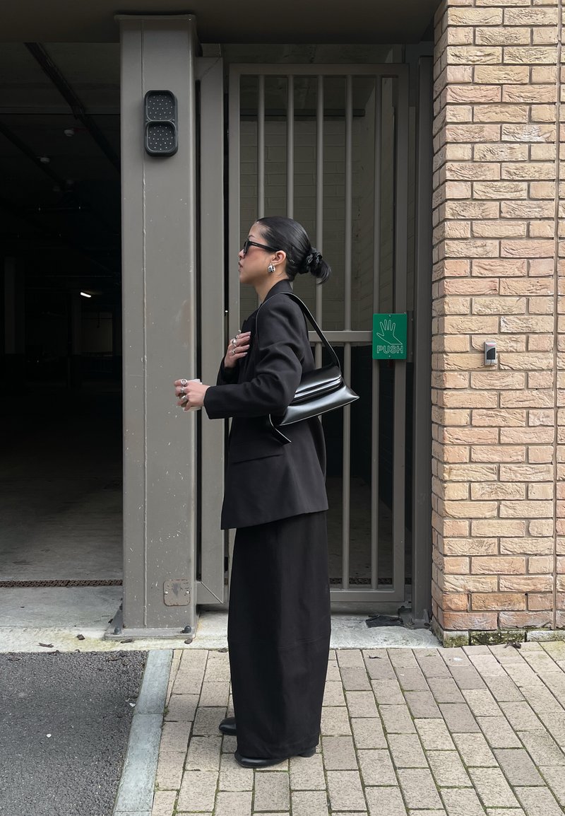 Γυναίκα με μαύρη ενδυμασία και γυαλιά ηλίου που κρατάει μαύρη τσάντα στέκεται δίπλα σε κλειστή μεταλλική πύλη με πράσινη ένδειξη "ΩΘΗΣΗ".