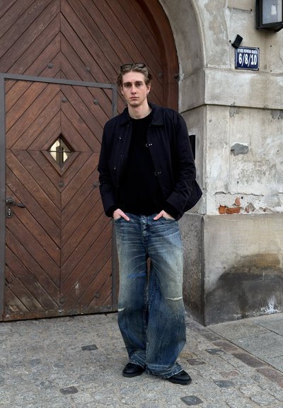 Joven con gafas en la cabeza, chaqueta negra y vaqueros anchos y desgastados, de pie con las manos en los bolsillos junto a una gran puerta arqueada de madera y una pared de piedra.