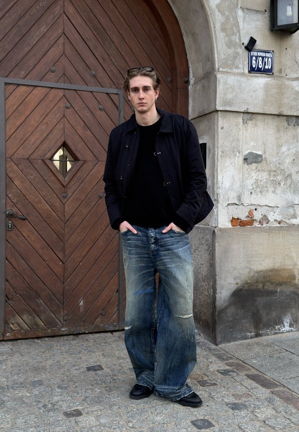 Jonge man met bril op het hoofd, zwarte jas en wijde versleten spijkerbroeken staand met handen in de zakken bij grote houten boogdeur en stenen muur.