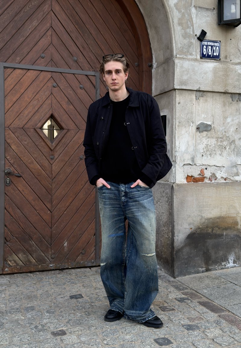 Jeune homme avec lunettes sur la tête, veste noire et jeans larges usés, debout les mains dans les poches près d'une grande porte en bois cintrée et d'un mur de pierre.