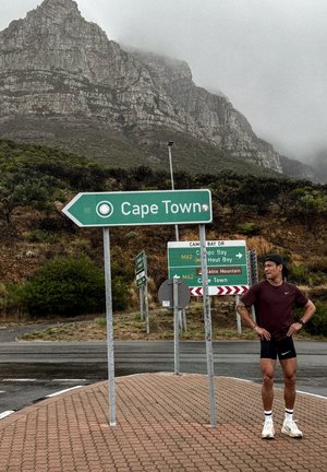 Corredor con ropa deportiva está junto a señales de tráfico que indican hacia Ciudad del Cabo y lugares cercanos en la base de una montaña cubierta de niebla.