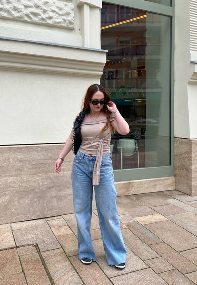 Top envolvente beige claro, jeans de pierna ancha de cintura alta en azul claro, gafas de sol negras y un bolso negro con acentos plateados, de pie cerca de una ventana de cristal.