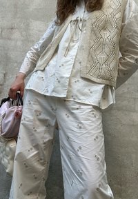 White cotton blouse adorned with floral embroidery, layered with a beige knitted vest. Loose-fitting white trousers showcase similar floral patterns.