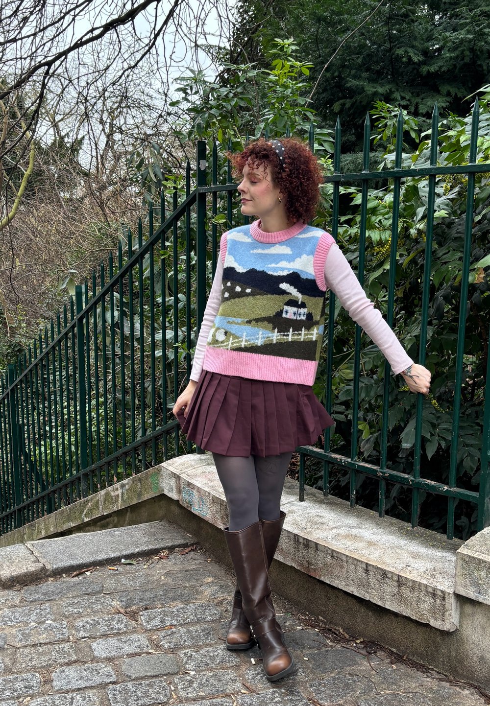 Jeune femme aux cheveux bouclés portant un gilet en tricot au motif pittoresque, une chemise rose à manches longues, une jupe bordeaux, des collants gris et des bottes marron, debout près d'une clôture verte.