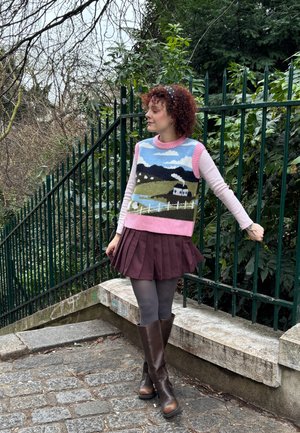 Joven con cabello rizado que lleva un chaleco tejido con paisaje, camisa de manga larga rosa, falda granate, medias grises y botas marrones, de pie junto a una cerca verde.