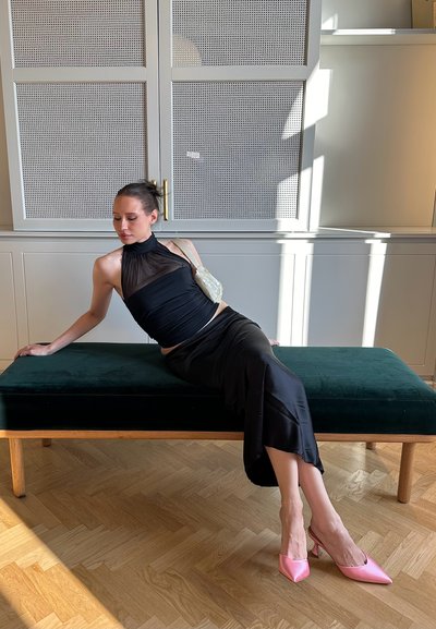 Top negro de satén con detalle fruncido y tirantes halter, combinado con una falda negra de satén. Tacones rosas de punta afilada. Sentada en un banco de terciopelo verde oscuro.