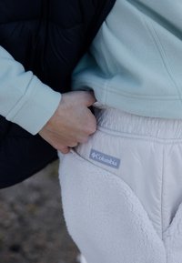 Camisola de polar azul claro combinada com calças de moletão brancas, com textura fofinha, apresentando uma etiqueta do logo da Columbia na cintura. Colete preto acolchoado visível.