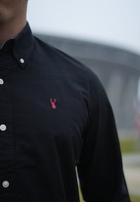 Chemise noire boutonnée en tissu à texture lisse, avec un petit logo de cerf rouge brodé sur la poitrine.