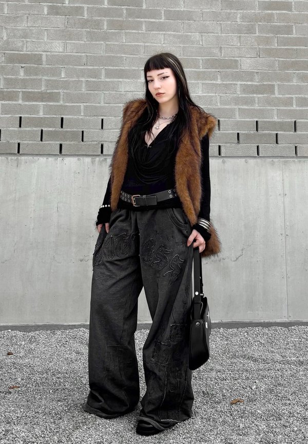 Jeune femme aux cheveux foncés portant une veste en fourrure, un haut noir, un pantalon noir large et tenant un sac à main noir contre un mur en briques.