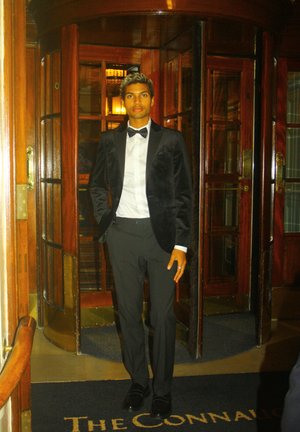 Chaqueta de esmoquin de terciopelo negro sobre una camisa blanca, pantalones negros y zapatos, de pie junto a una puerta giratoria en un interior elegante.