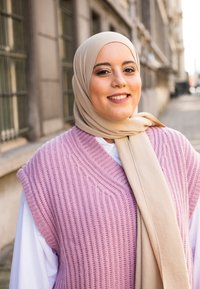 Colete de malha rosa claro com um design de decote em V, sobreposto a uma camisa branca; hijab bege drapeia elegantemente. Textura suave e ajuste casual.