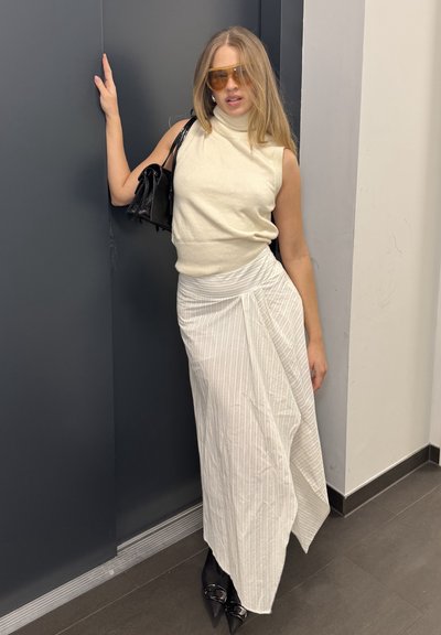 Top sin mangas de punto en color blanco roto, falda blanca con rayas drapeada, zapatos de tacón negros y bolso negro. Se llevan gafas de sol con cristales amarillos.