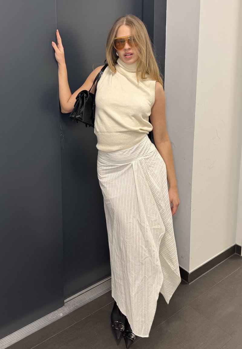 Haut sans manches en tricot de couleur blanc cassé, jupe blanche drapée à rayures, chaussures noires à talons et sac à main noir. Lunettes de soleil teintées jaune portées.