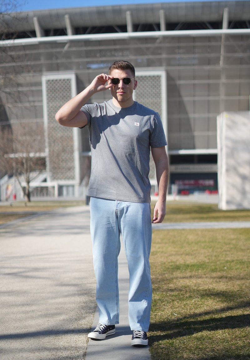 Jeune homme portant des lunettes de soleil, un t-shirt gris, un jean bleu clair et des baskets, debout sur un chemin près d'un bâtiment moderne en plein jour.