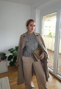 Manteau trench en beige, pull en tricot texturé gris avec des boutons dorés, pantalon large beige et écharpe pliée. Tenant un sac à main en brun avec des détails en chaîne.
