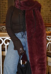 Personne en jean bleu et haut marron à manches longues portant des gants foncés, tenant un sac à main noir, avec de longues tresses et une grande écharpe bordeaux à l'intérieur.