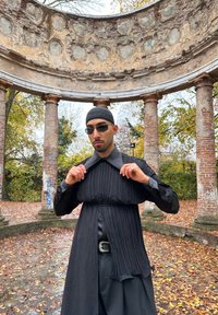 Homme portant une chemise noire plissée, des lunettes de soleil et une casquette, ajustant son col sous un pavillon circulaire en pierre, entouré de feuilles d'automne tombées.