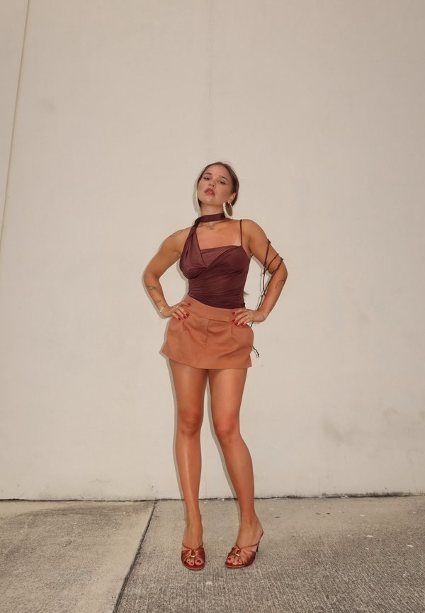 Femme posant les mains sur les hanches, portant un haut asymétrique marron rouge, une mini-jupe beige, des boucles d'oreilles anneaux et des talons bruns devant un mur uni.