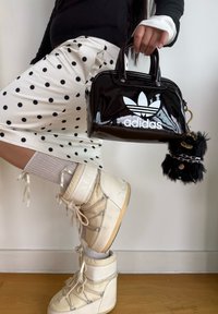 Čierna lakovaná kabelka s bielym logom Adidas, strieborným hardwarom a nadýchaným príveskom; kombinovaná s bielymi šortkami s bodkami a bielymi čižmami.