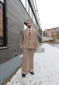 Abito marrone a quadri con pantaloni a gamba larga e grandi rever, abbinato a una cravatta marrone. Scarpe nere e sfondo neutro con neve.