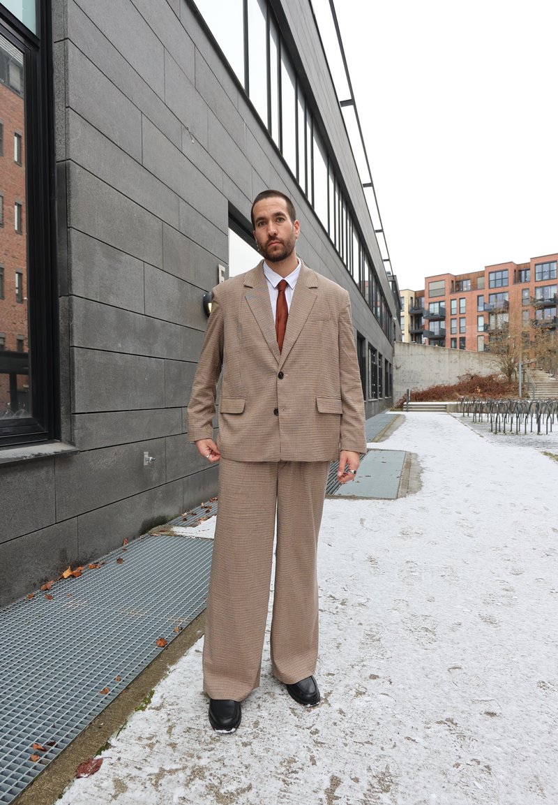 Costume marron à carreaux avec jambes larges et grands revers, assorti d'une cravate marron. Chaussures noires sur un fond neutre avec de la neige.