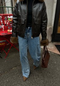 Personne portant une veste en cuir marron foncé avec des poignets en fourrure, un jean bleu clair, des chaussures pointues marron, tenant un sac à main marron sur un trottoir en ville.