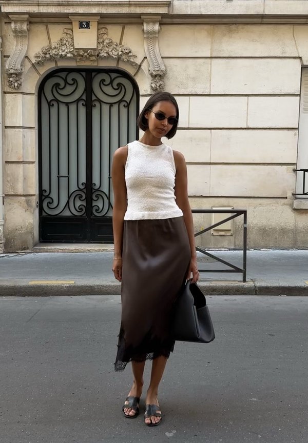 Mujer con camiseta blanca sin mangas, falda oscura, gafas de sol negras y sandalias, de pie en una calle de la ciudad sosteniendo un bolso negro.