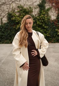 Kremowy, oversize'owy płaszcz z teksturowanej tkaniny, noszony na dopasowanej brązowej sukience. Długie włosy, minimalna biżuteria oraz brązowa torebka.