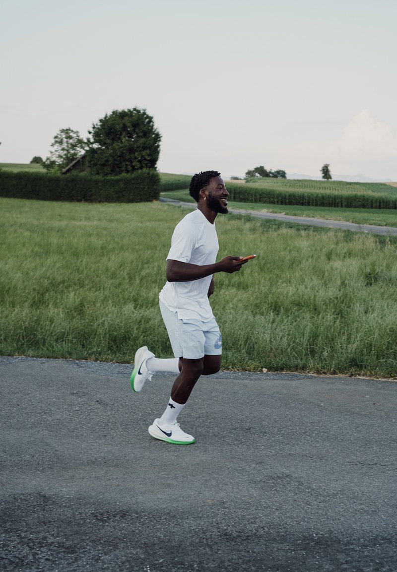 Homme en tenue de sport blanche faisant du jogging sur une route rurale en tenant un smartphone, avec des champs verts et des arbres en arrière-plan.
