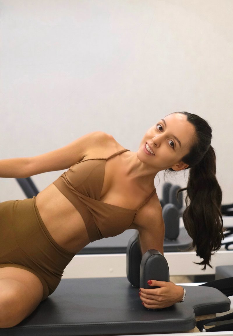 Jeune femme en tenue de sport marron fait de l'exercice sur un appareil de Pilates reformer, souriante, avec ses longs cheveux attachés en queue de cheval.