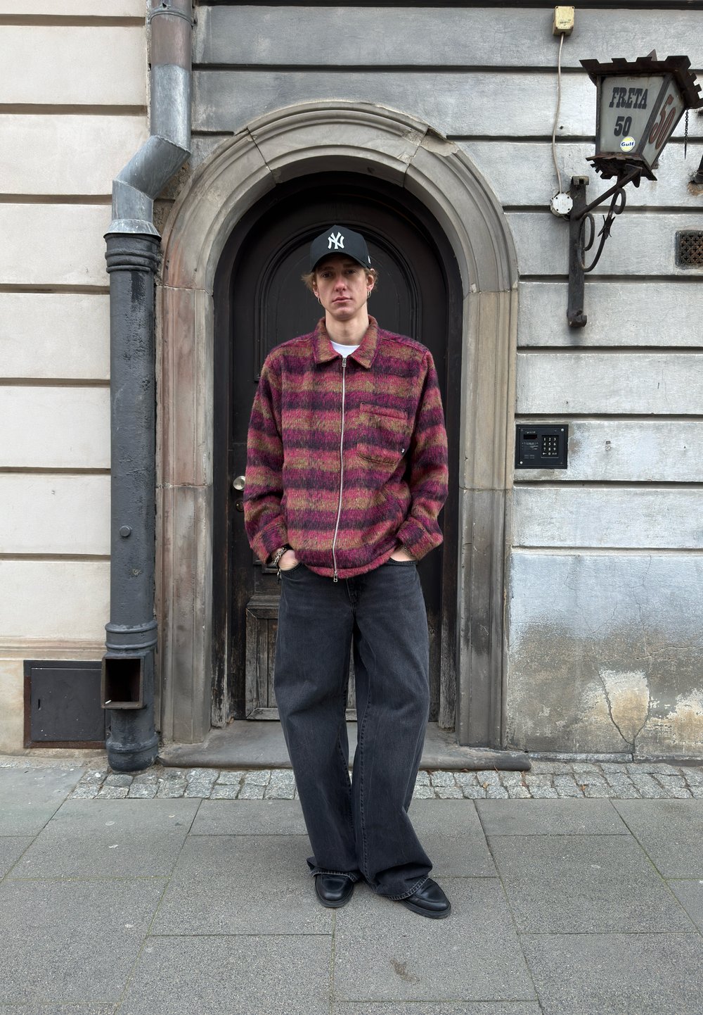 Jeune homme portant une casquette noire et une veste rouge à motifs, les mains dans les poches, debout devant une porte voûtée en bois sombre dans une rue pavée.