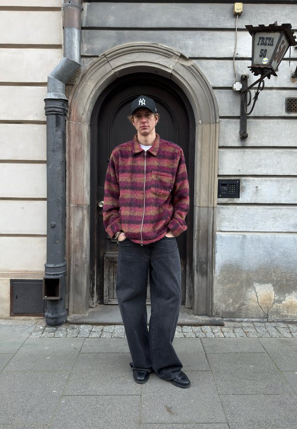 Young man wearing a black cap and a patterned red jacket stands with his hands in his pockets in front of an arched dark wooden door on a stone-paved street.