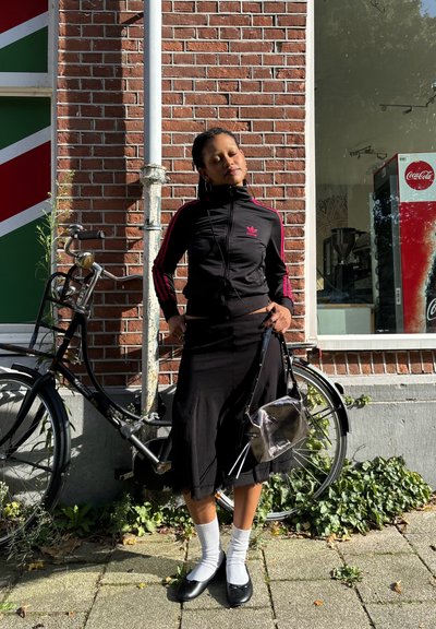 Chaqueta negra con cremallera y franjas rosas, falda negra, calcetines blancos y bailarinas negras. Sosteniendo un bolso transparente junto a una bicicleta.