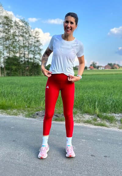 Leggings atléticos rojos con logo blanco y una camiseta corta blanca. Presenta zapatillas con acentos en rosa claro y blanco. Entorno exterior con fondo verde.