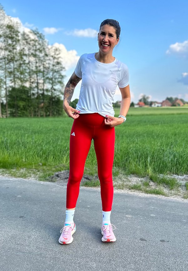 Czerwone legginsy sportowe z białym logo oraz krótka biała koszulka. W zestawie sneakersy w jasnoróżowe i białe akcenty. Plenerowe otoczenie z zielonym tłem.