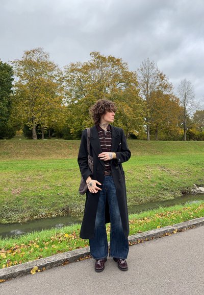 Abrigo largo negro sobre una camiseta a rayas, jeans anchos azules y zapatos burdeos. Sosteniendo una bolsa marrón, de pie al aire libre con árboles de fondo.