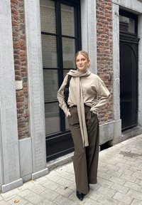 Beige gestrickter Pullover mit einem langen Schal, kombiniert mit hoch taillierten braunen weitem Hosen und spitzen schwarzen Absätzen, vor einer Ziegelwand.