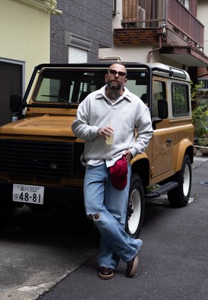 Hombre con tatuajes y gafas de sol se apoya en un Land Rover color mostaza, sosteniendo una bebida y una gorra roja, en un callejón urbano.