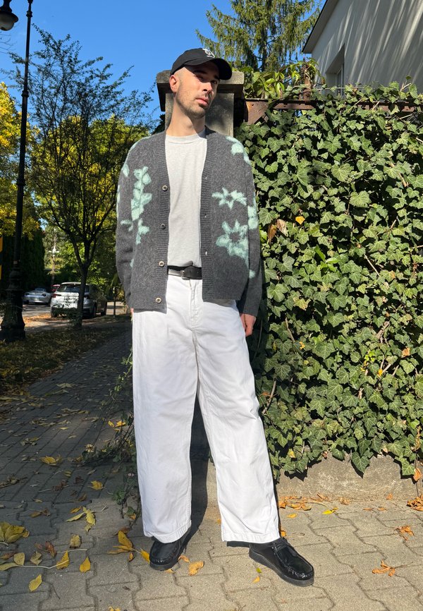 Homme debout à l'extérieur à côté d'une clôture recouverte de lierre, portant une casquette noire, un cardigan gris à motifs, un pantalon blanc et des chaussures noires par une journée ensoleillée.
