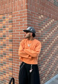 Oranje cropped lange mouwen shirt met witte logo, zwarte wijde broek met kettingdetail, en een grijze pet met een ontwerp, staand tegen een bakstenen muur.