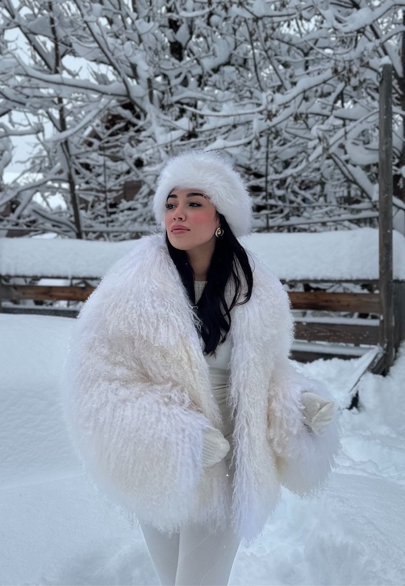 Woman wearing a white fluffy coat and hat stands outdoors in snow with snow-covered trees and wooden fence in the background.