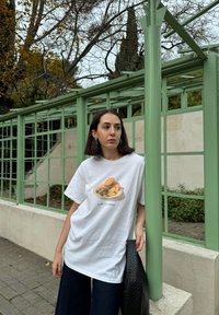 Hvid t-shirt med et grafisk motiv af småkager og bær, parret med mørke vide bukser og en struktureret sort taske mod en grøn rutenet baggrund.