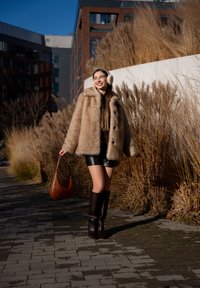 Femme en manteau de fourrure beige, short noir et bottes, marchant sur un chemin pavé, portant un sac à main marron avec de hautes herbes et des bâtiments en arrière-plan.