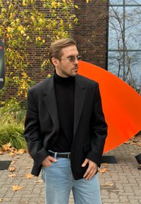 Blazer nero sopra un dolcevita nero, abbinato a jeans azzurri. Accessori includono occhiali sottili con lenti colorate. Lo sfondo presenta arancioni e vegetazione.