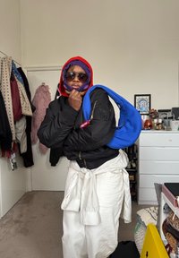 Personne portant un couvre-chef coloré superposé, des lunettes de soleil, une veste noire, un pantalon blanc noué à la taille, tenant un sac à main bleu sur l'épaule dans une chambre.